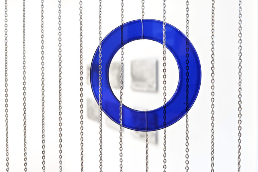 Ein blauer, gläserner Ring hängt zwischen mehreren vertikalen Metallketten vor weißem Hintergrund.