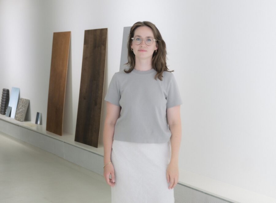 Eine Frau mit Brille, grauem T-Shirt und weißem Rock steht vor Holzpaneelen an einer weißen Wand.