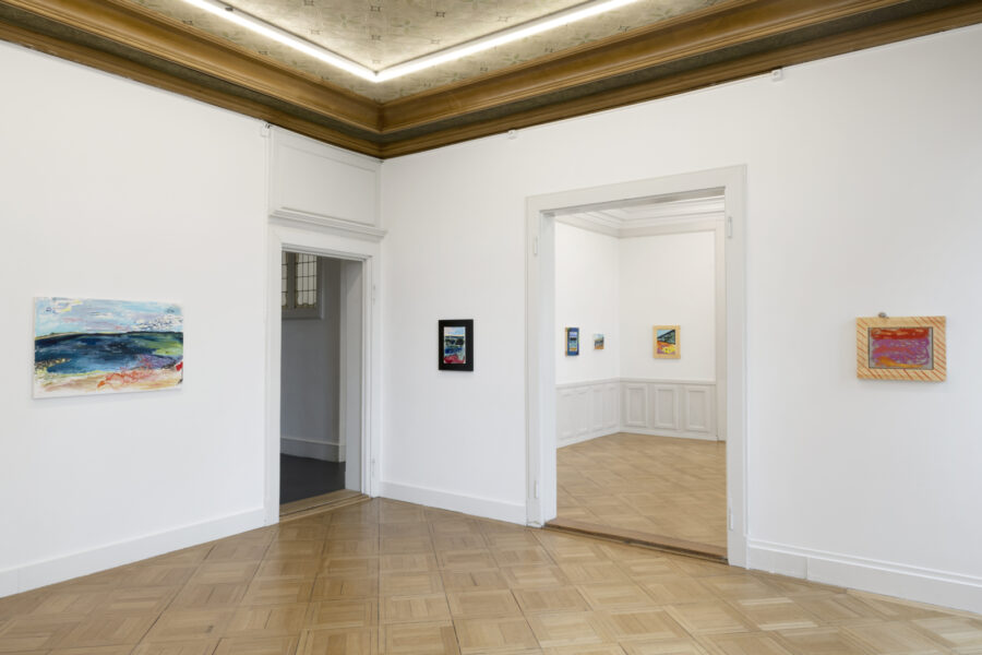Helle Galerie mit weißem Parkettboden und mehreren farbigen Gemälden an den Wänden.