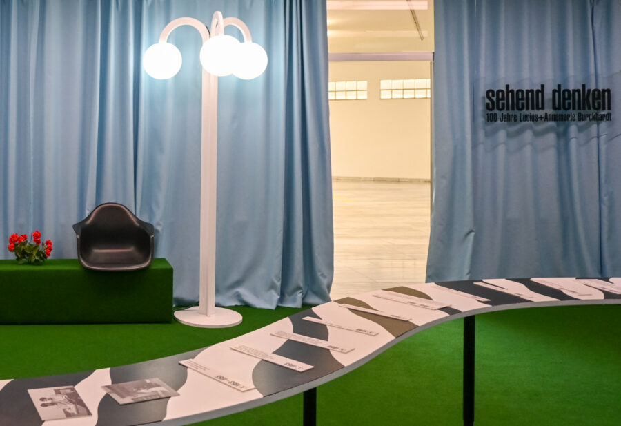 Ausstellungsraum mit blauen Vorhängen, weißer Stehlampe, schwarzem Sessel und geschwungener Tafel.