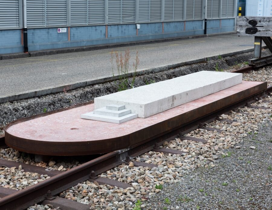 Eine weiße, rechteckige Steinskulptur auf einer rostigen Metallplattform über Bahngleisen.