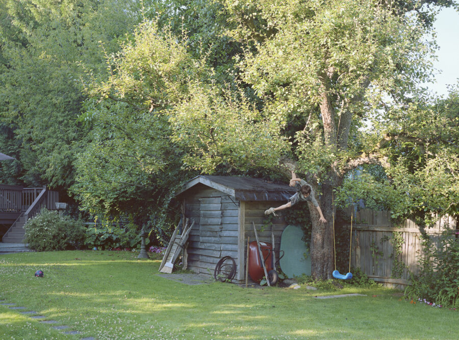 Das Foto zeigt eine Gartenszenerie. Auf einer grünen Gartenfläche steht ein Gartenhäuschen aus Holz. Daneben steht ein Baum, in dem eine blaue Schaukel baumelt. Ein Junge in kurzen Hosen fällt gerade vom Baum herunter.