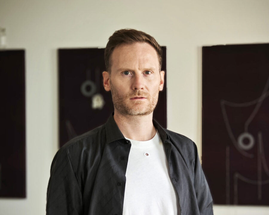 Porträt eines bärtigen Mannes in einem schwarzen Hemd über einem weißen T-Shirt vor Kunstwerken.
