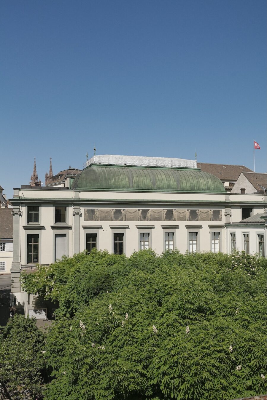 A white neoclassical building with a green copper roof, partially obscured by lush green trees.