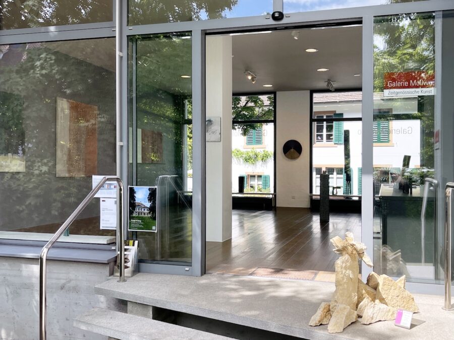 Glass entrance to Galerie Mollert, showing a modern art gallery interior and an outdoor sculpture.