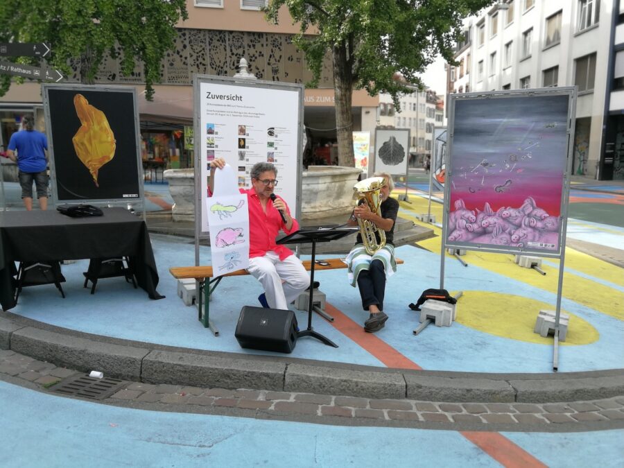 A man speaks into a microphone while a musician plays a tuba at an outdoor art exhibition.