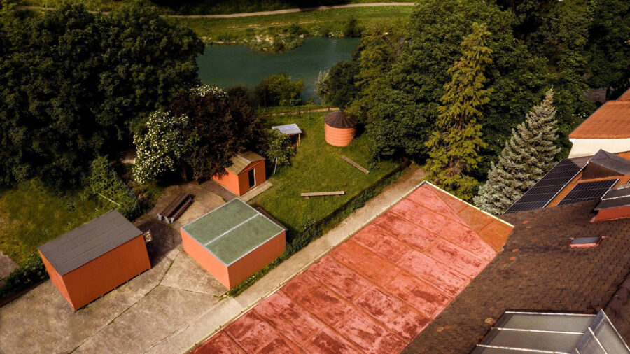 Aerial view of a yard with small orange sheds, a lawn, and a river surrounded by green trees.