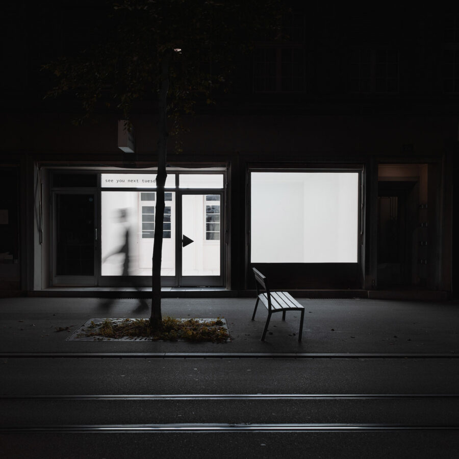 A blurred figure walks past a lit storefront with a white sign and an empty bench at night.