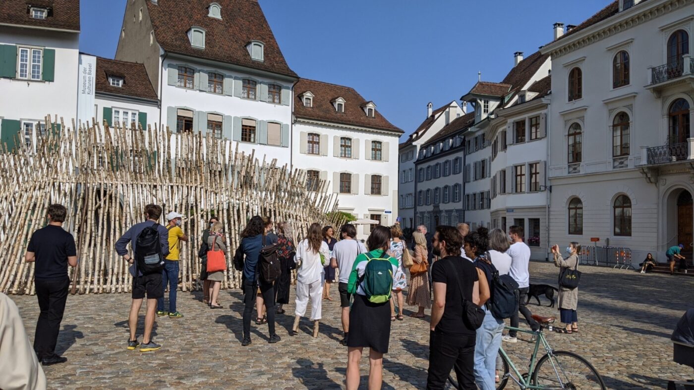 Eine Gruppe von Menschen betrachtet eine große Holzstelen-Installation auf einem Stadtplatz.