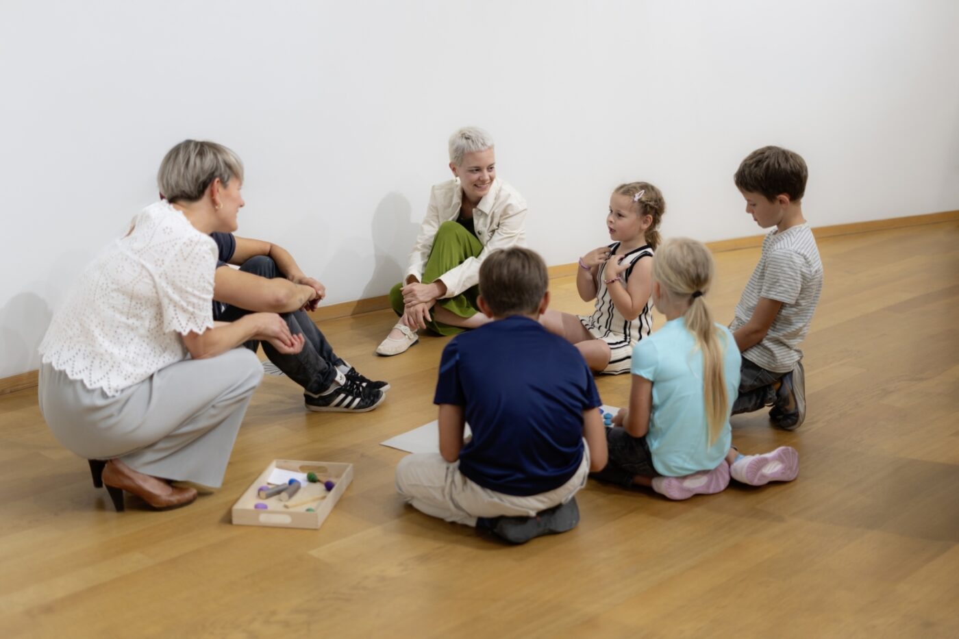Zwei Erwachsene und drei Kinder sitzen in einem hellen Raum im Kreis auf dem Holzboden.