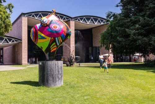 Eine bunte, abstrakte Skulptur auf einer grünen Wiese vor einem modernen Gebäude.