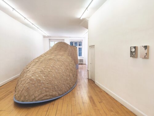 A large, woven fabric art installation fills a narrow white gallery with wooden floors.