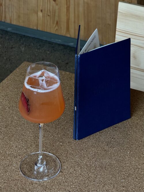 Ein orangefarbener Cocktail im Glas und eine dunkelblaue Speisekarte auf einem Korktisch.
