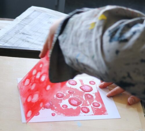 Eine Person hebt ein rot gefärbtes Blatt Papier von einem künstlerischen Druck ab.