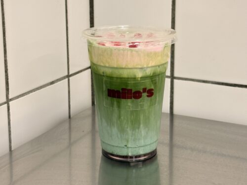 Ein grüner Schichtdrink im Plastikbecher mit dem Logo „milo's“ vor einer weißen Kachelwand.