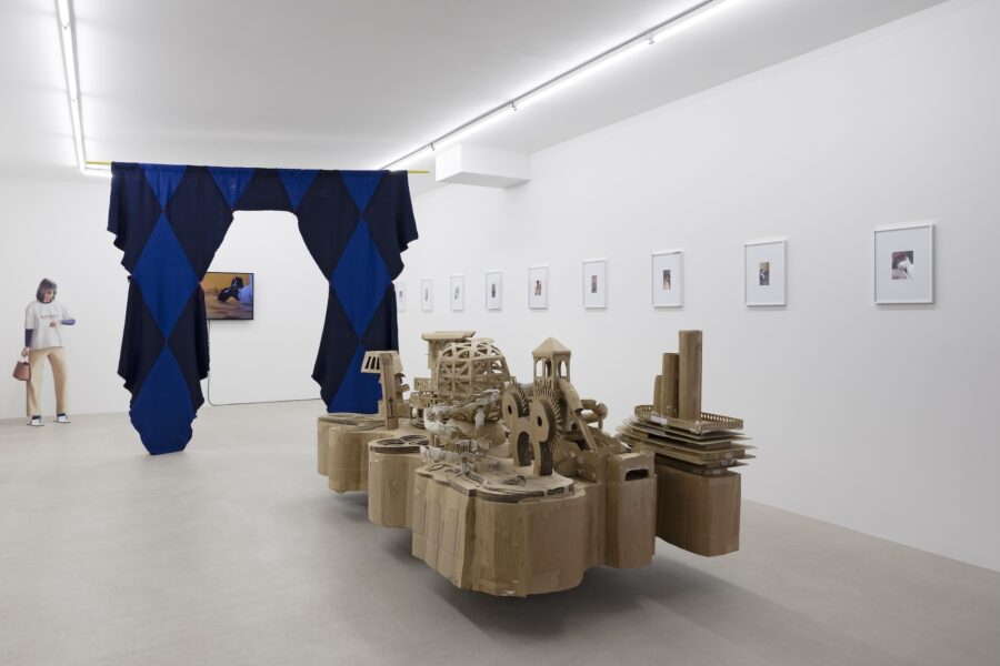 Kunstausstellung mit einer Pappskulptur, einem blau-schwarzen Vorhang und Bildern an der Wand.