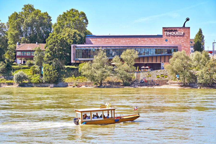 A small tour boat cruises on a river in front of the Museum Tinguely and lush green trees.