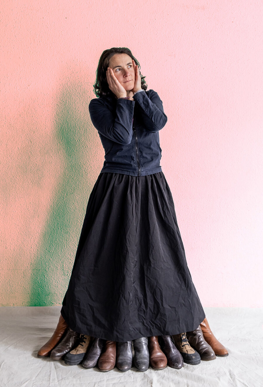 A woman in a black skirt and jacket poses with her hands on her face, surrounded by many shoes.