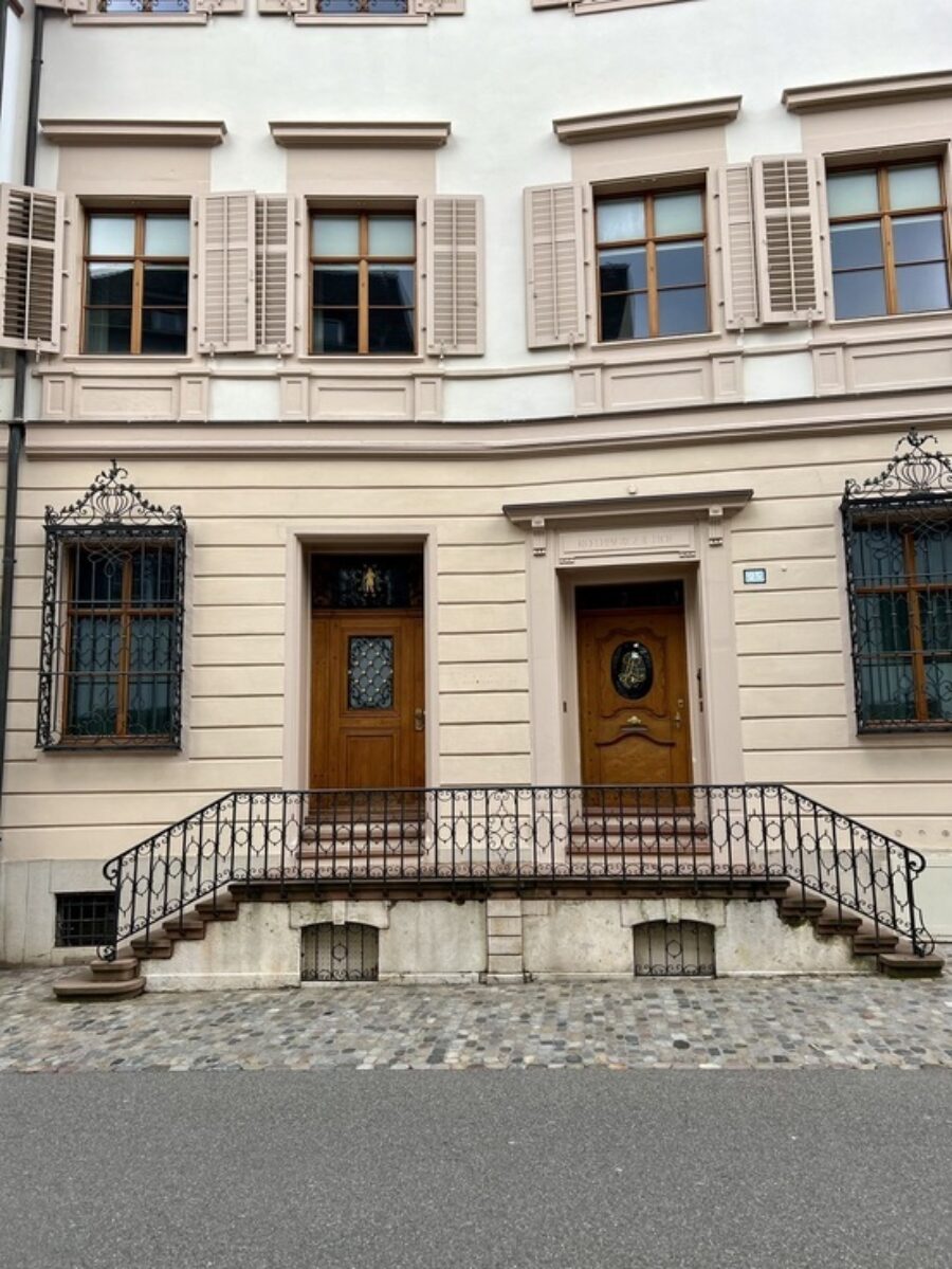 Facade of a light-colored historic building with two wooden doors and black wrought-iron railings.