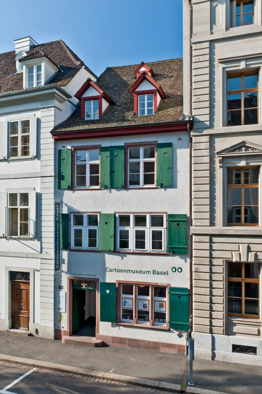 Facade of the Cartoonmuseum Basel, a narrow white building with green shutters and a brown roof.