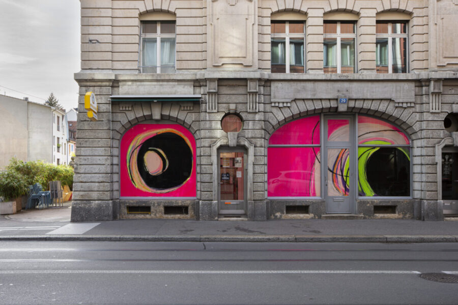 Storefront of a stone building with vibrant pink and yellow abstract art in the windows.