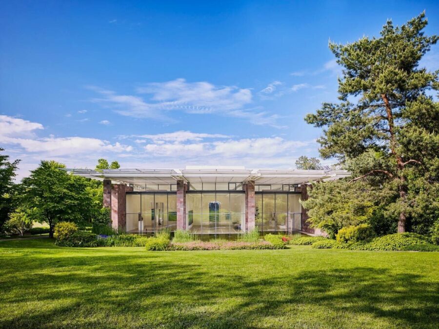 Modernes, gläsernes Gebäude mit flachem Dach inmitten einer grünen Parklandschaft unter blauem Himmel.