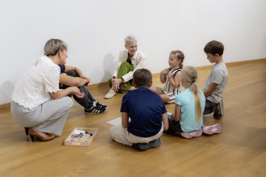 Two women and three children sit in a circle on a wooden floor in a white room.