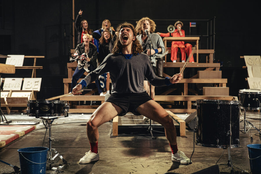 Eine Band performt energisch auf einer Bühne mit hölzernen Podesten und Schlagzeugen.