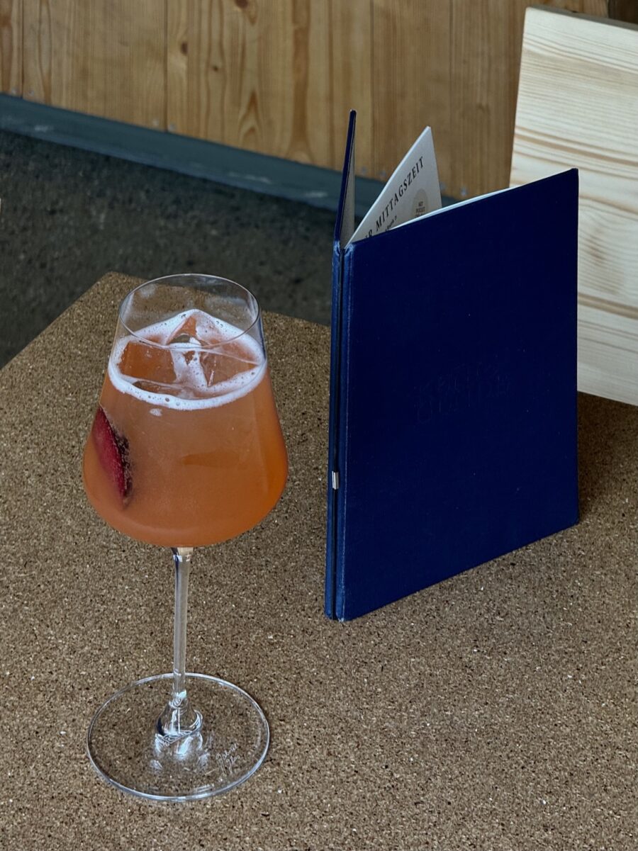 Ein orangefarbener Cocktail im Glas und eine dunkelblaue Speisekarte auf einem Korktisch.