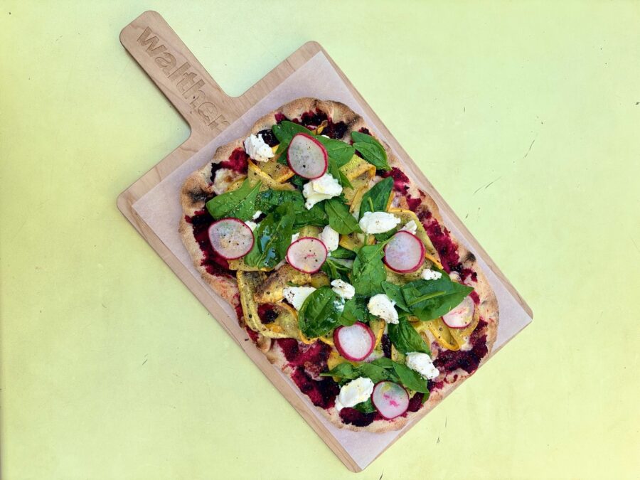 Eine längliche Pizza mit Rote-Bete-Basis, gelbem Belag, Mozzarella, Spinat und Radieschen auf Holz.
