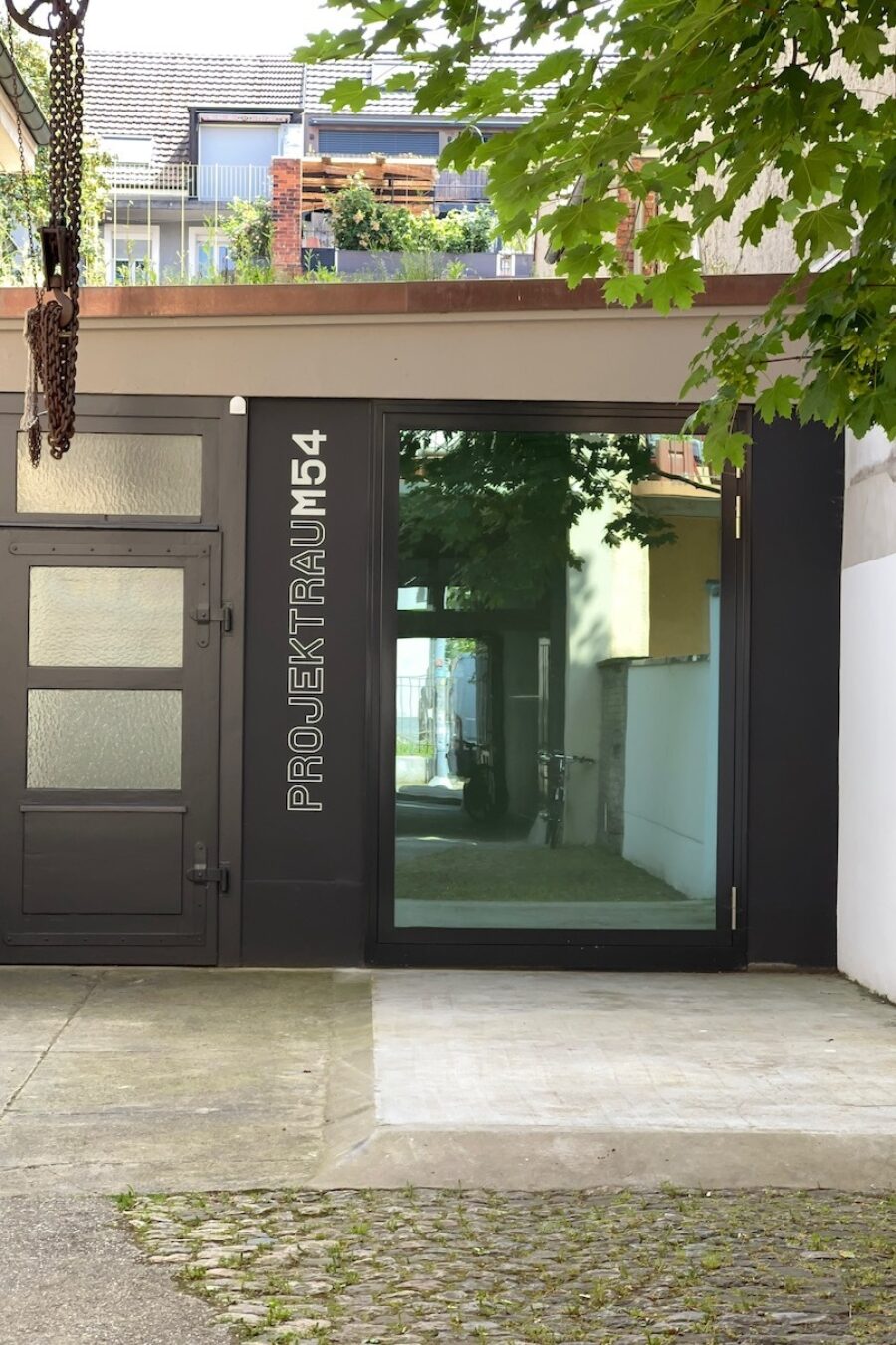 Entrance to Projektraum 54 with a dark gray door, a glass window, and green foliage above.