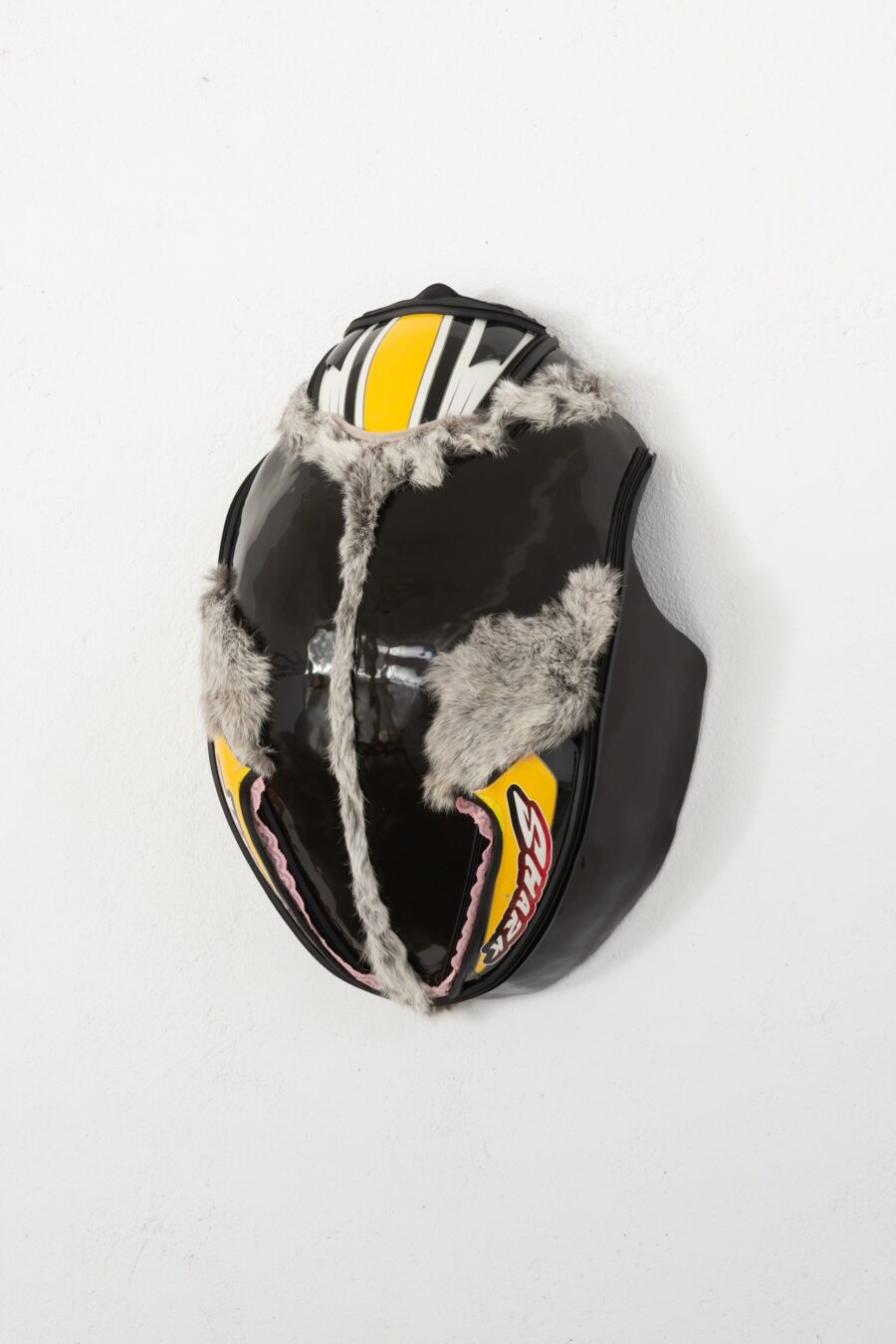 Ein schwarzer, glänzender Helm mit grauem Kunstfell und gelben Akzenten vor weißem Hintergrund.