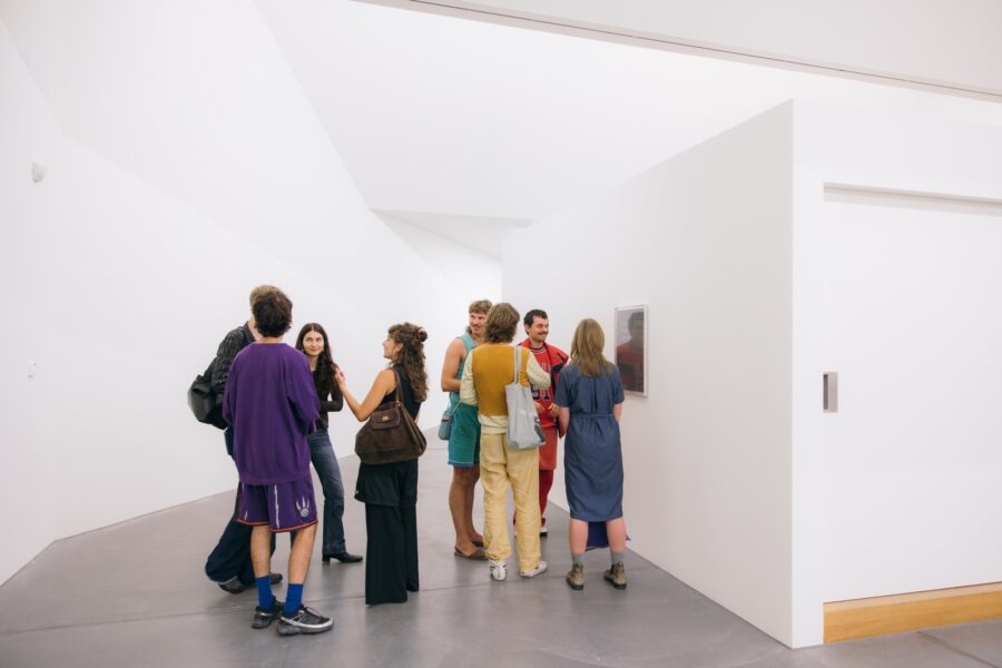 Eine Gruppe von Menschen steht in einem hellen Galeriebereich vor einem Kunstwerk an der Wand.