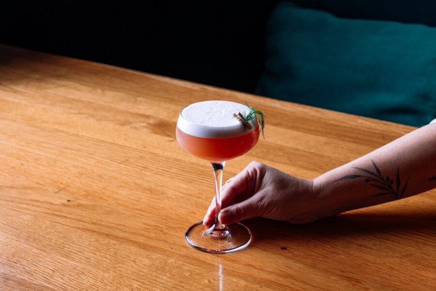 A hand holds a red cocktail with a white foam top and a rosemary garnish on a wooden table.