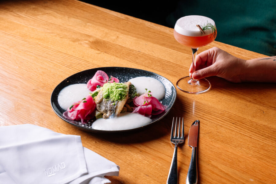 A plated fish dish with pink pickled vegetables and a hand holding a cocktail on a wooden table.