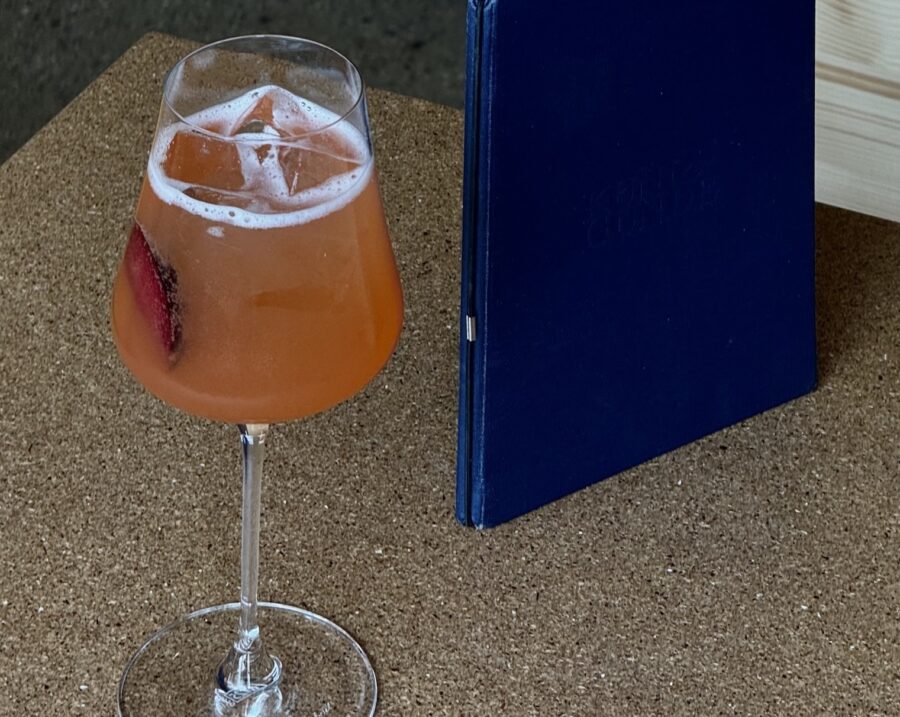 An orange-colored cocktail with foam and ice in a wine glass next to a blue menu on a cork table.