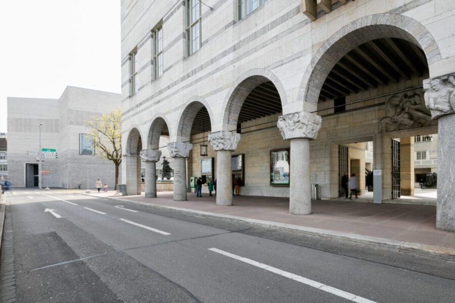 A stone building with a series of large arches and decorative columns along a city street.