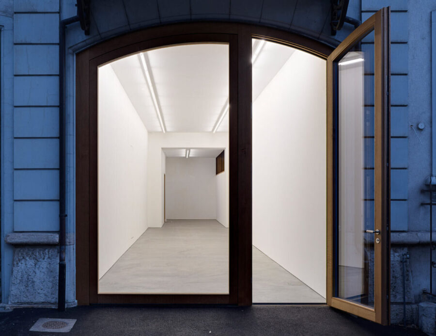 View through an open arched wooden doorway into a bright, empty white art gallery.