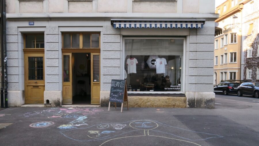 Storefront of a creative studio with white t-shirts in the window and chalk art on the sidewalk.