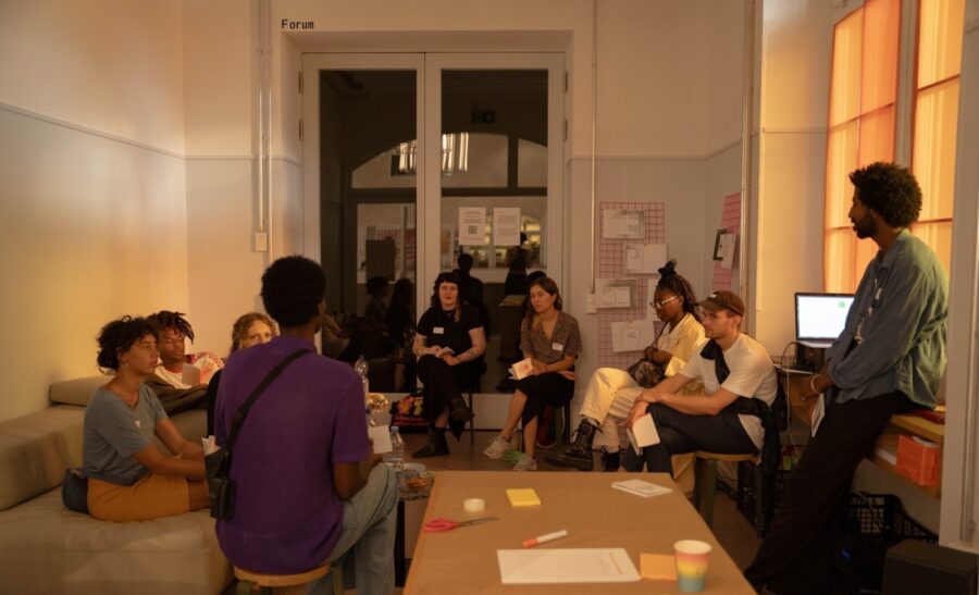 Das Foto zeigt einen Raum in gelblichem Licht in dem 11 Personen in einem Kreis sitzen und sich zugewandt sind, sich austauschen. Links ist ein Sofa zu sehen und in der Mitte ist ein Tisch, auf dem Notizen niedergeschrieben sind.
