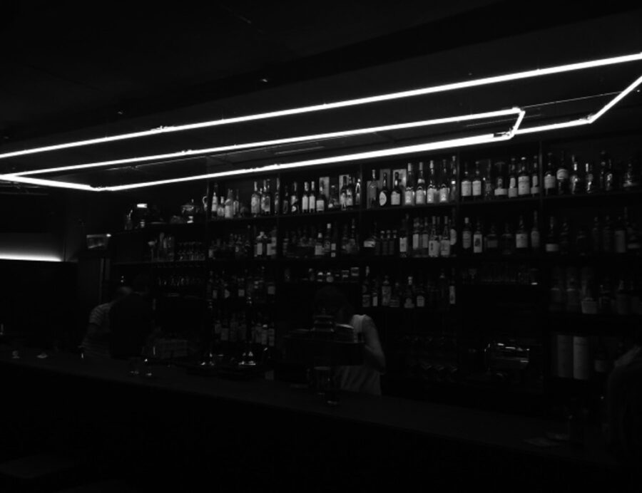 Black and white photo of a dark bar with illuminated linear lights over shelves of bottles.